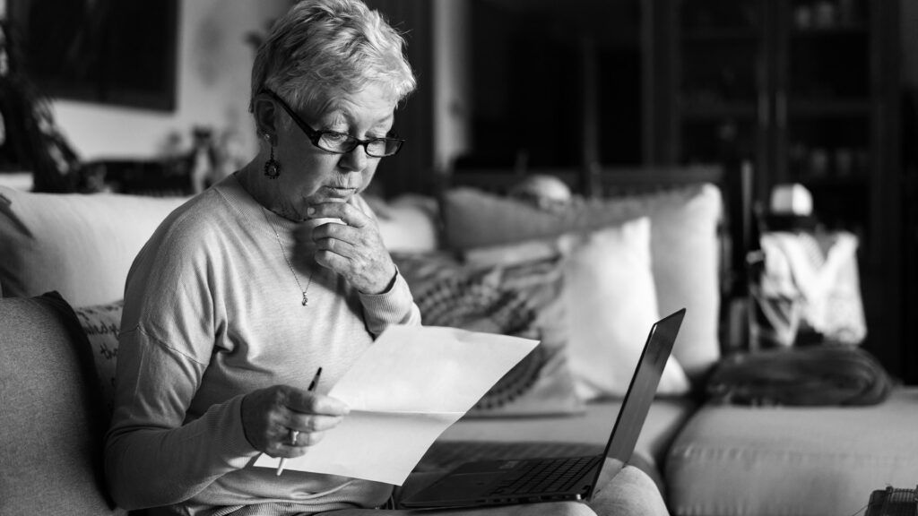 Older female adult sitting on a couch with a laptop on her knees holding a piece of paper and a pen