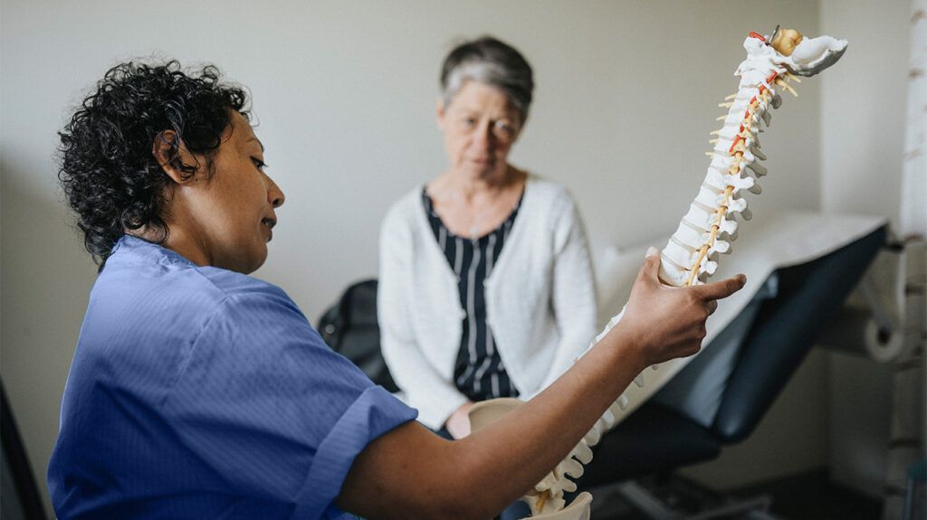 Nurse and patient in a medical setting. The nurse is holding up a model of the spine and pointing something out