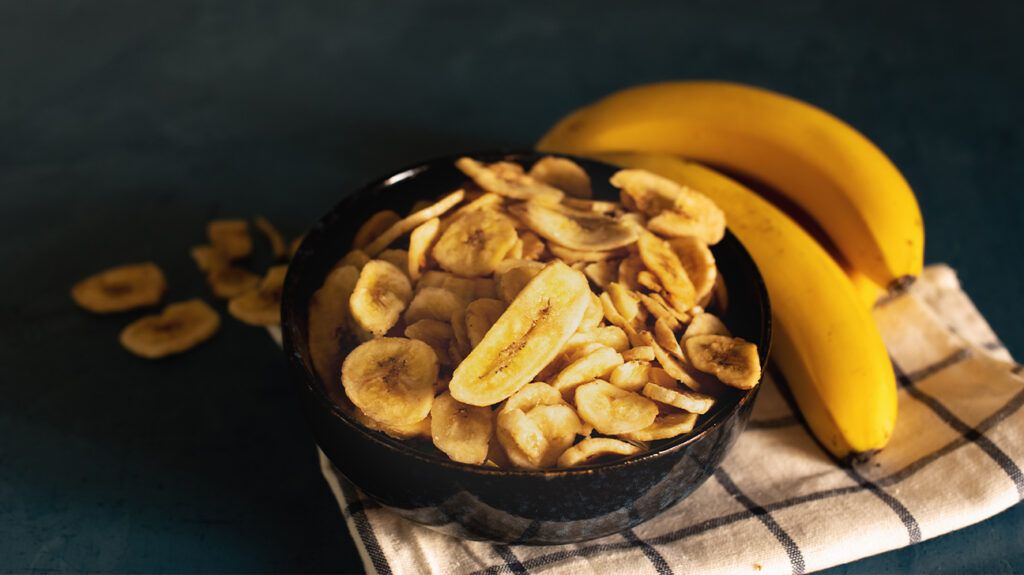 A bowl of dried banana slices next to two whole bananas.-1