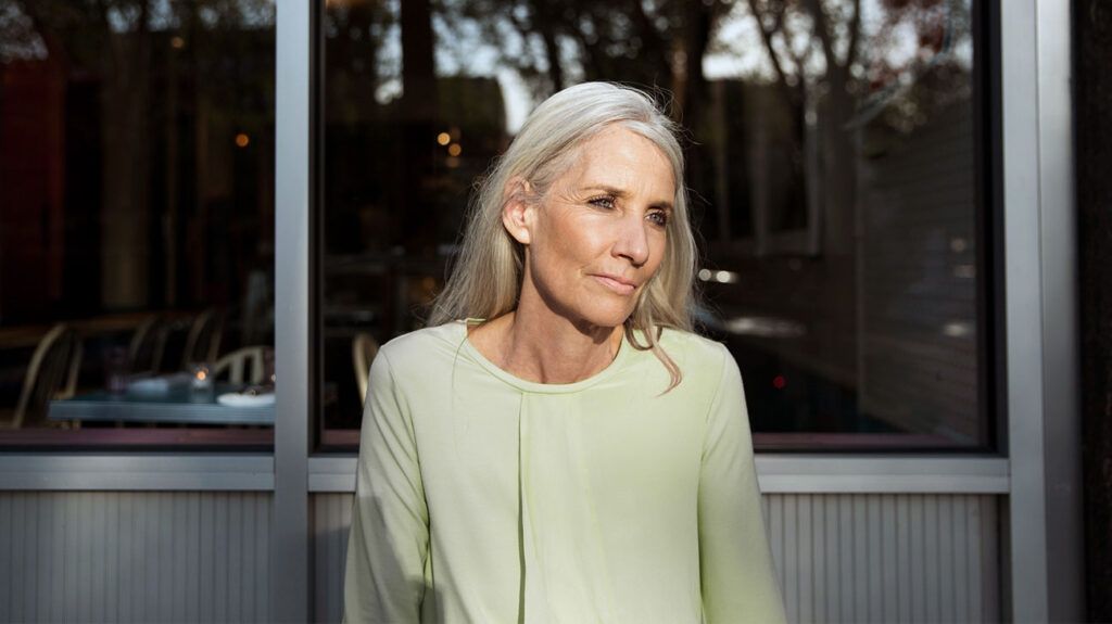 An older adult woman looks slightly to the side, outside as the sun shines on her in front of a glass panel building