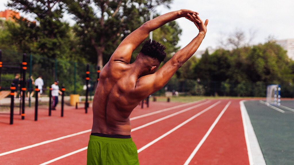 An athlete stretching by doing a side bend while stood before a running track.