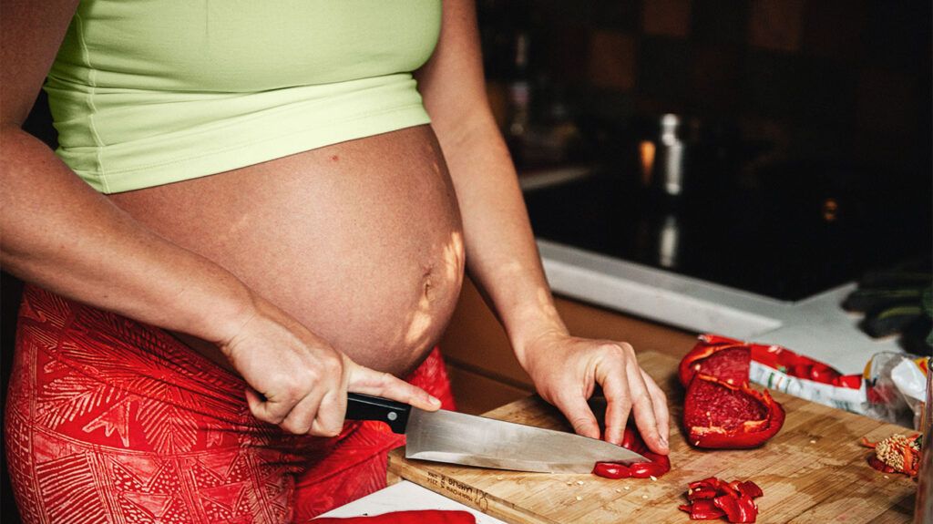 A heavily pregnant person chops peppers on a chopping board in a kitchen