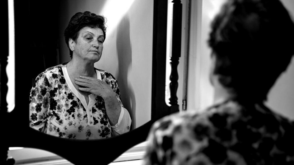 Black and white photo of an older woman looking in the mirror with her hand on her chest