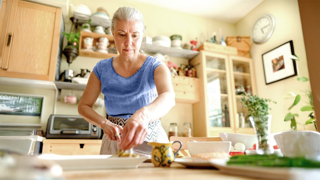 An older adult woman preparing a Mediterranean salad in a kitchen with a knife, chopping board and various utensils