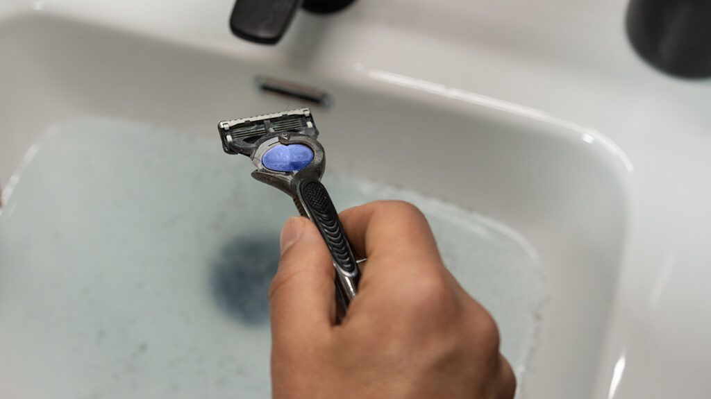 A hand holding a razor above a sink full of water. -1