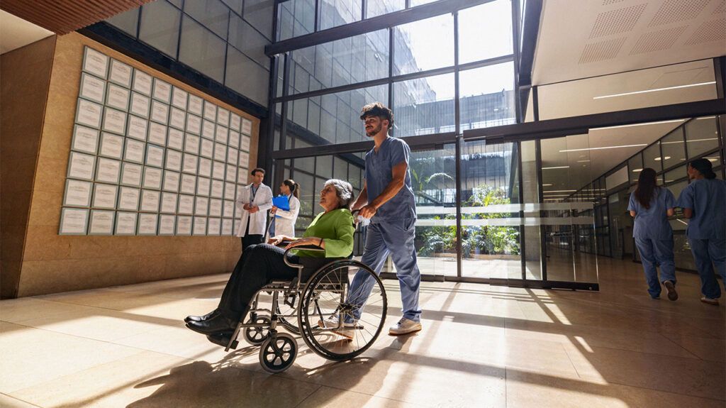 A nurse pushing an older adult in a wheelchair through a hospital atrium made of glass.