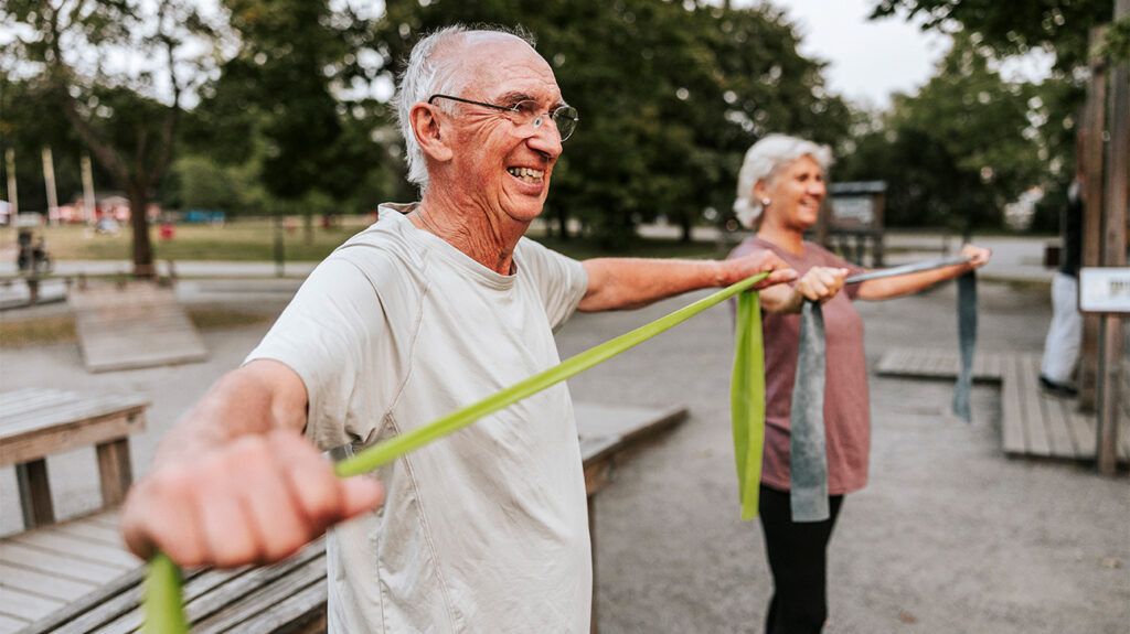 Two older adults doing arm band stretching exercises outside in a park