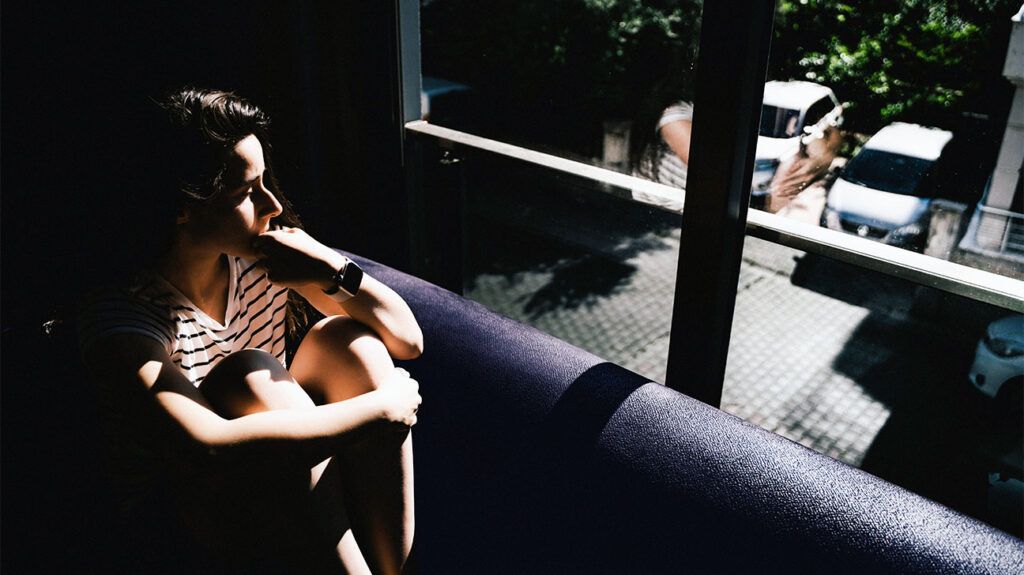 A person sitting, clutching knees to chest, looking out of a window.