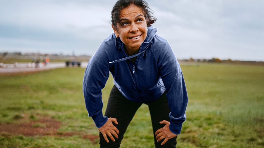 A middle aged woman holds her knees, her body slightly bent over, catching her breath after a run outside
