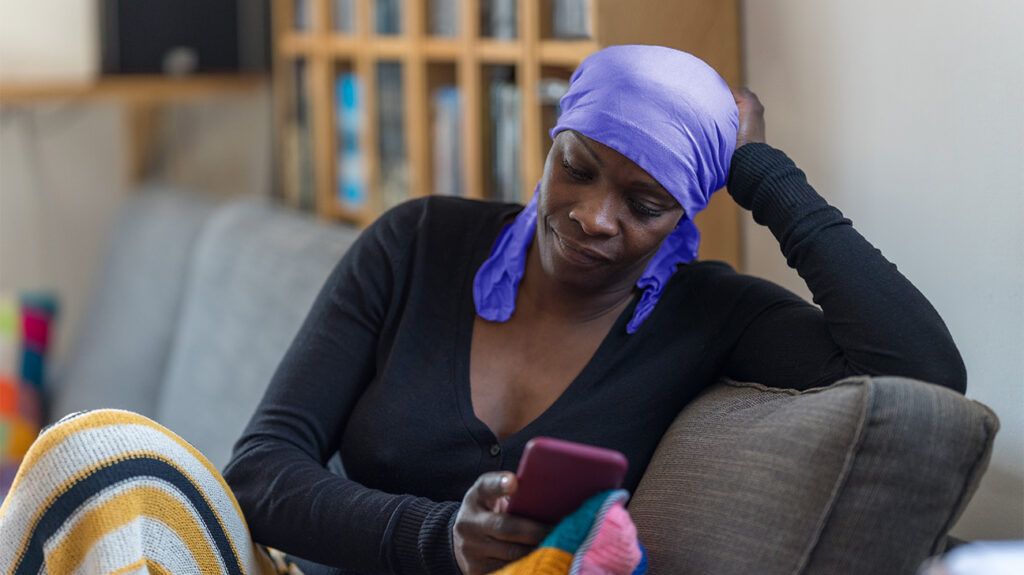 Black woman wearing purple head scarf and black shirt sits on the sofa looking at her smartphone