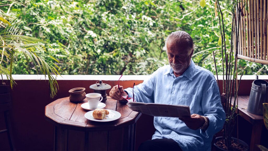 An older adult reads a newspaper with his glasses in hand as he has a breakfast of coffee and a croissant next to a table near a window