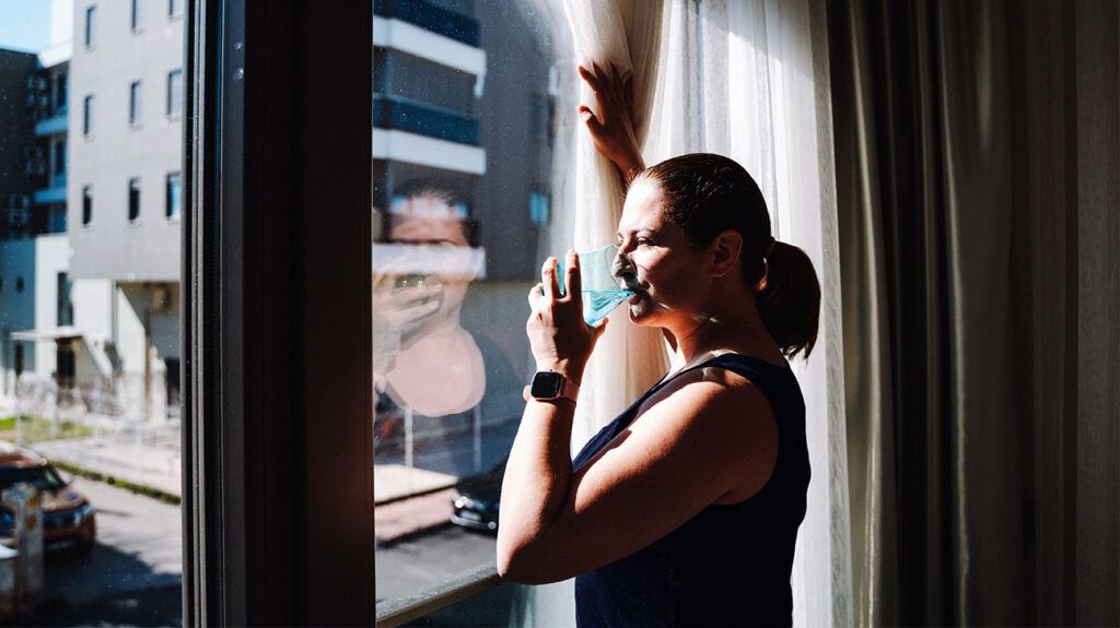 A person drinking a glass of water in the morning while looking out of their window.