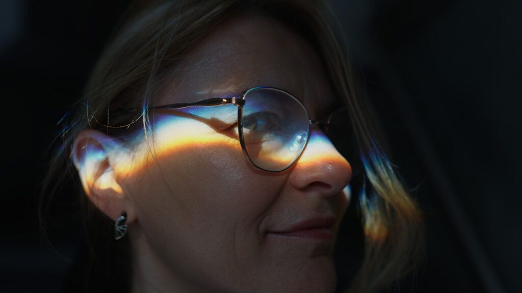 A prism of light and rainbow colours shine on a woman's face wearing glasses