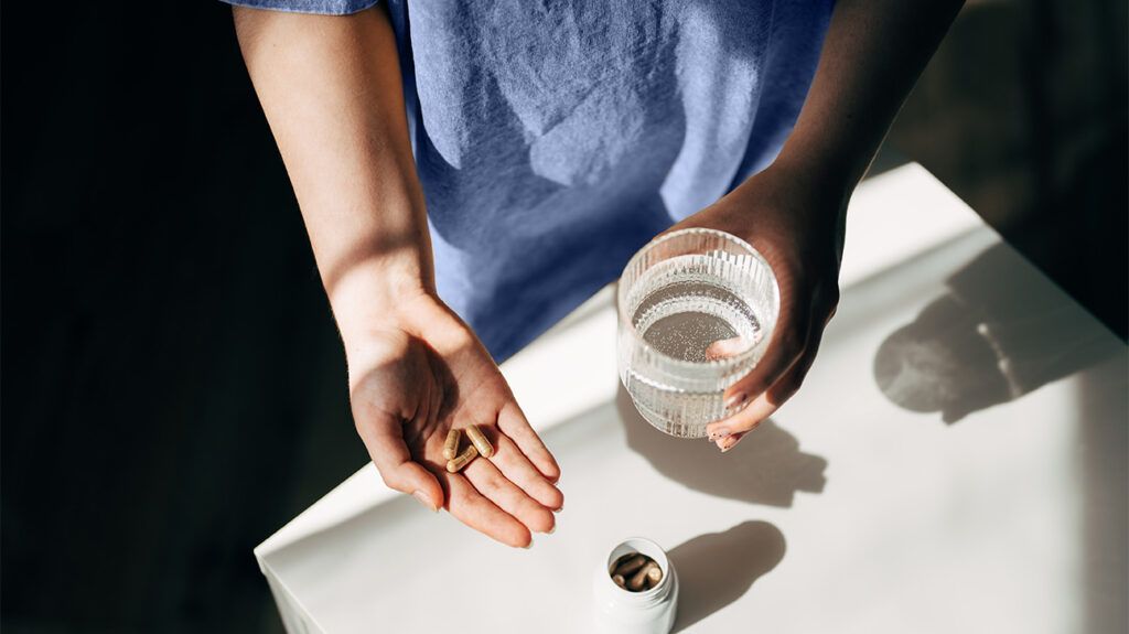 A person holding a few vitamin D capsules in one hand and a glass of water in another over a table