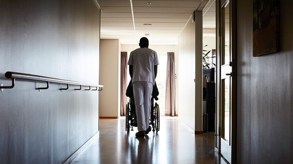 Black nurse in white slack wheeling patient down hospital corridor