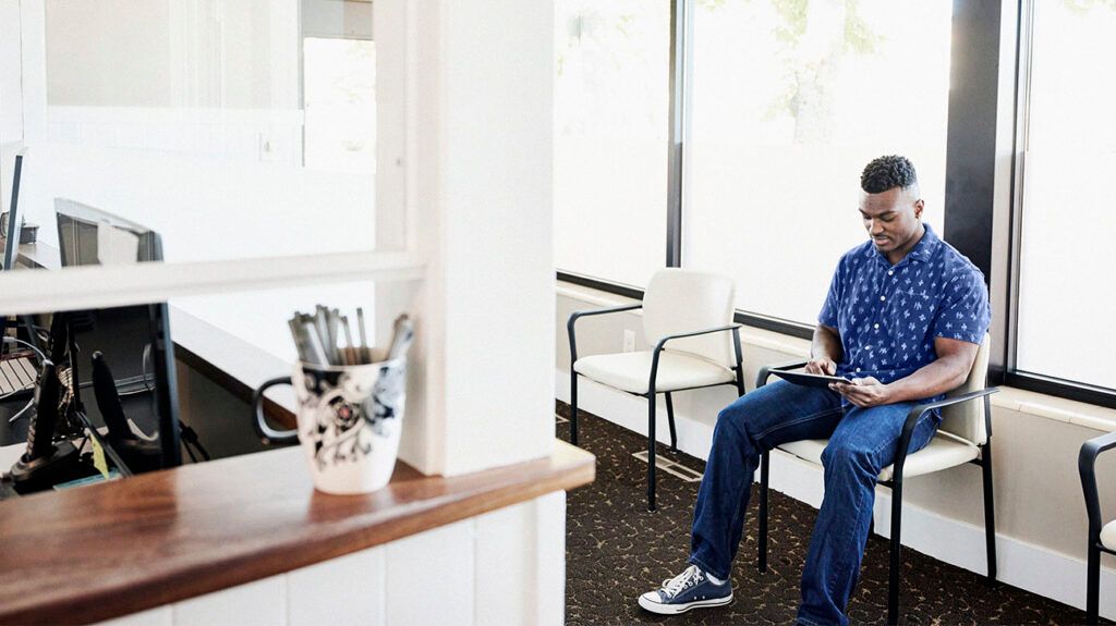 Wide shot man in medical waiting room completing form on digital tablet