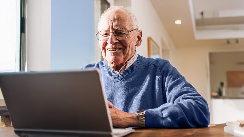 An older adult male works on a laptop, smiling at the screen