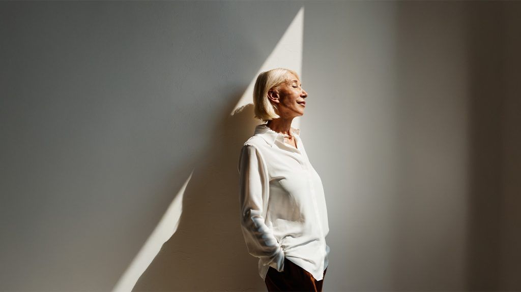 An older woman stands with her body slightly angled sideways against a wall as a ray of light illuminates her face