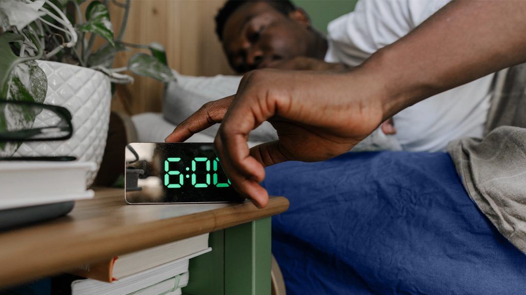 A man presses a digital alarm clock to turn it off while laying in bed