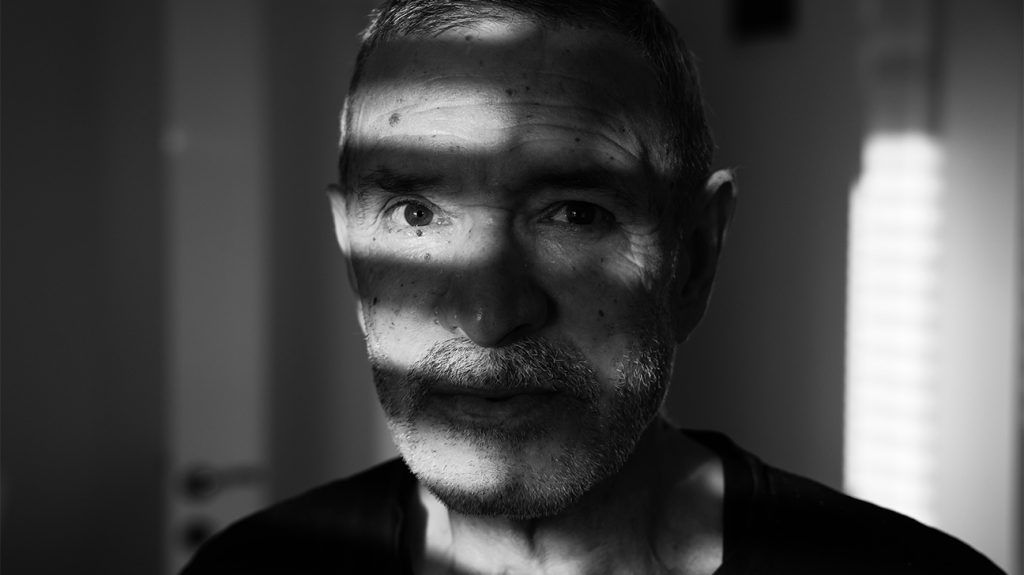 An older adult looking straight ahead as light from window blinds casts shadows on their face.