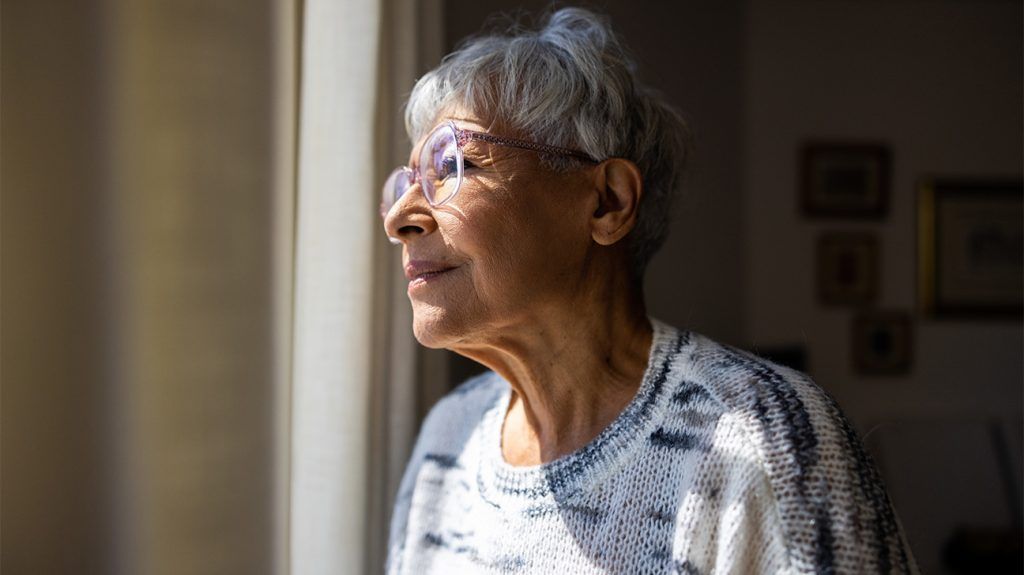 An older adult woman looks out of the window pensively sunlight shining on half her face