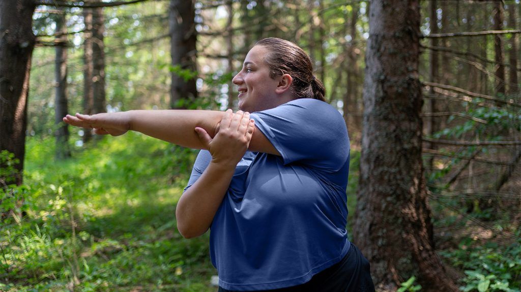 A woman with central obesity stretches her arms in the woods as part of her workout