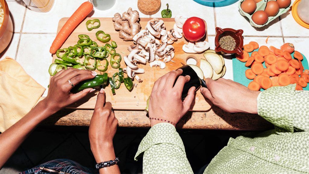 Two people cut various vegetables on a wooden chopping board side by side