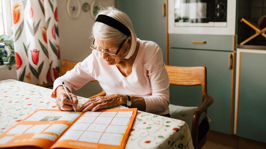 나이 든 백인 여성이 부엌 식탁에서 십자말풀이를 풀고 있다.<BR>