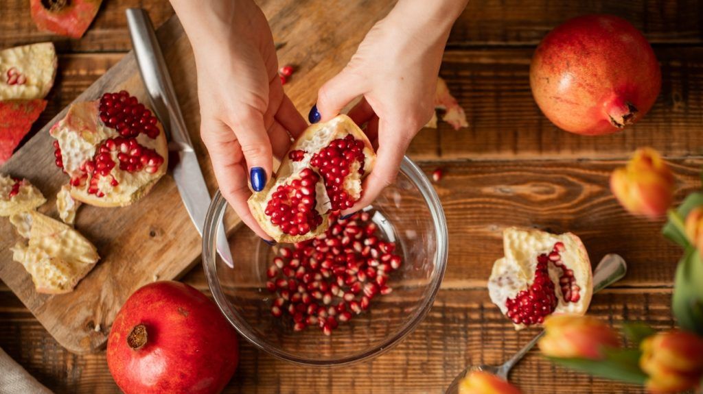 hands holding pomegranate above pomegranate seeds in a bowl