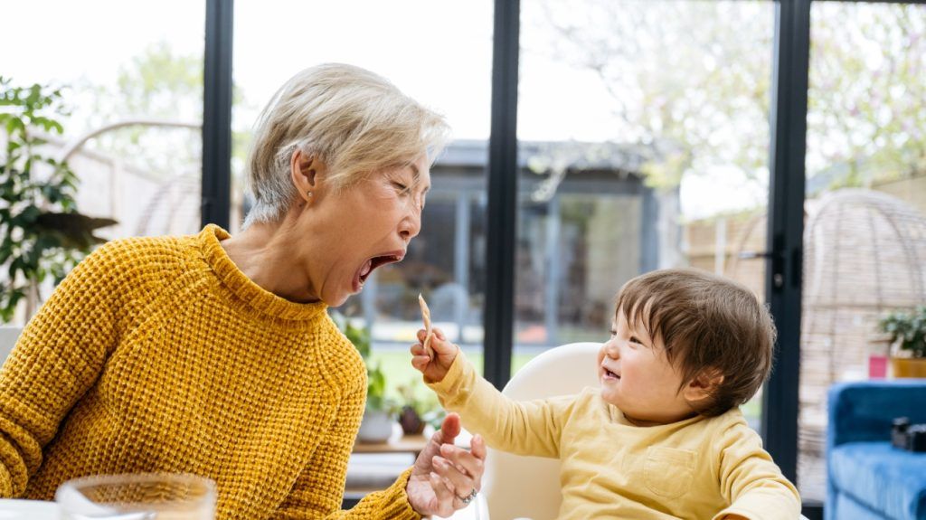 A toddler feeds their grandmother at a table with a spoon while sitting in a high chair at a dinner table