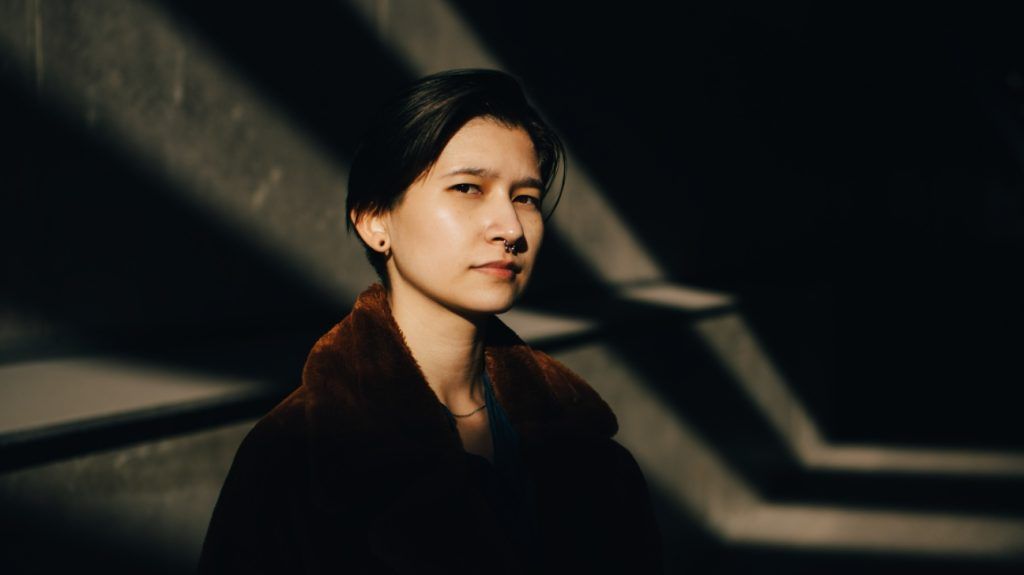 Portrait of young person with cropped black hair and nose piercing standing against wall