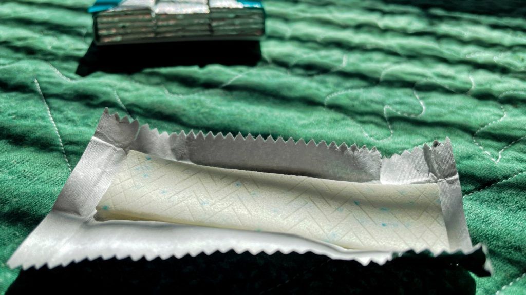 An opened stick of chewing gum contianign artificial sweeteners lays on a flat surface in front of a full pack.