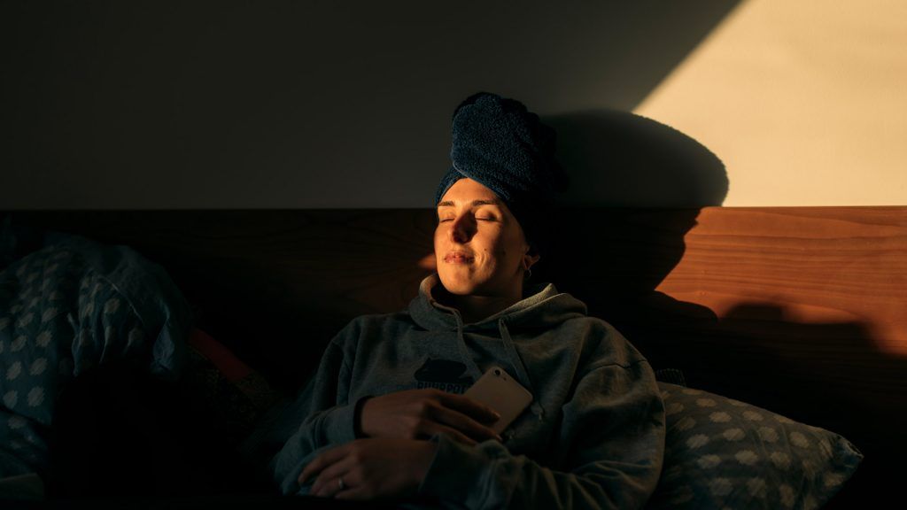 Woman resting in bed with her phone, her eyes are closed, light from a window is shining on her face