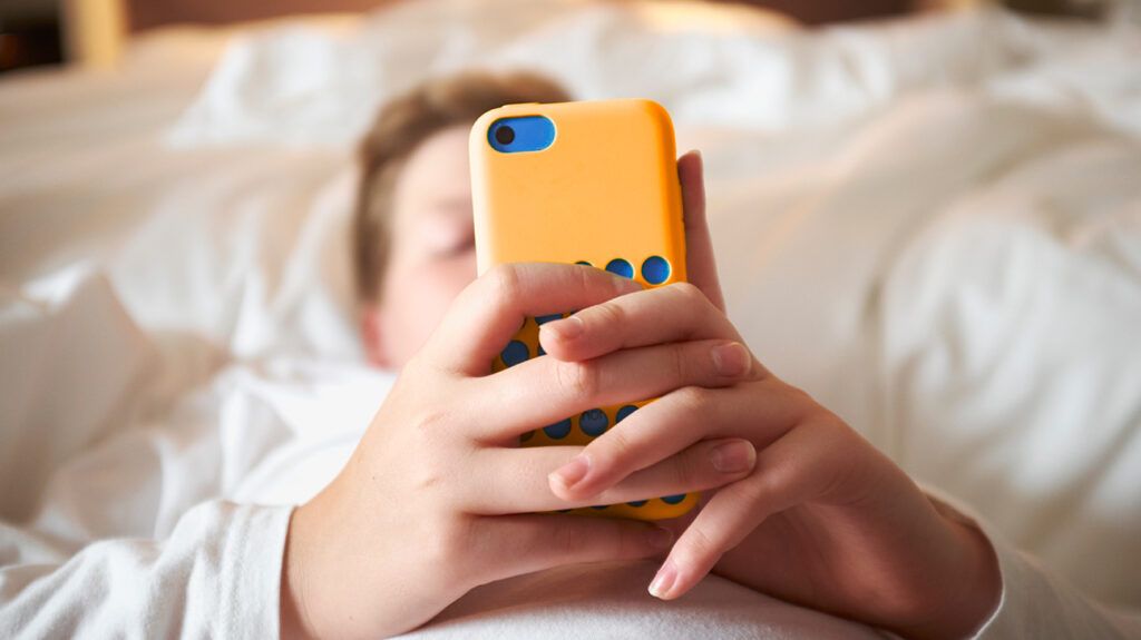 Close up of a teenage boy lying in bed holding a yellow smart phone