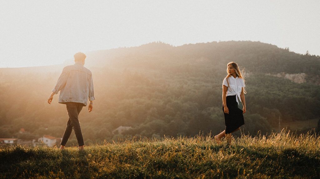 Couple walking outdoors a bit of distance between them. They look at each other. The sun is slowly setting.