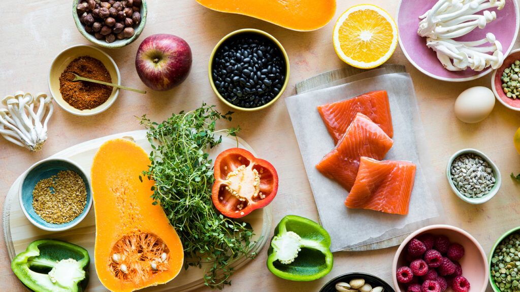 An assortment of brightly colored and healthy foods, including butternut squash, salmon, bell pepper, beans, nuts, seeds, and berries