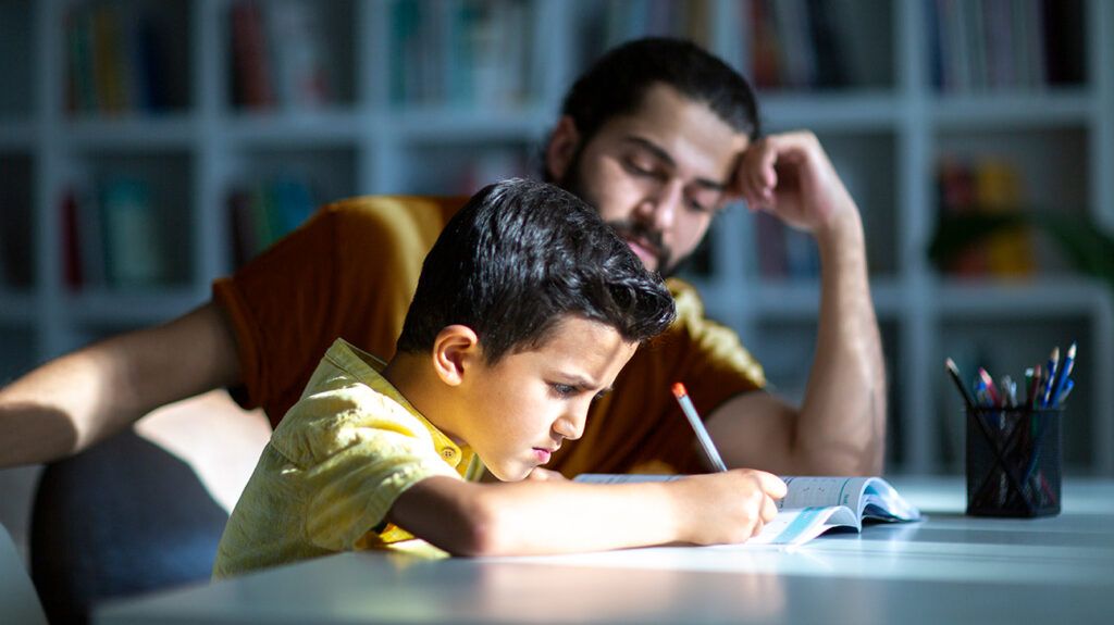 A boy doing homework at a table with the father observing and helping him
