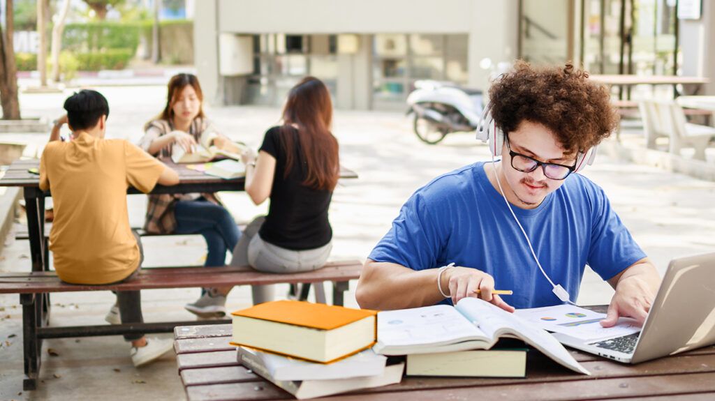 A college student studying and reading book in an outdoor campus location, behind him on a different table three students are chatting