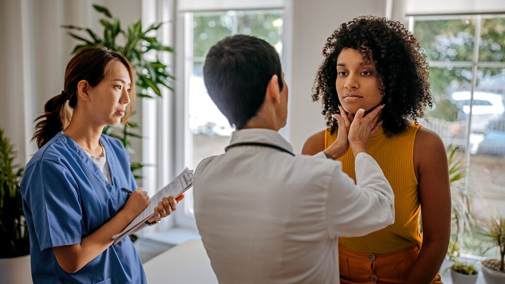 A female doctor doing a thyroid exam on a female patient with a nurse taking notes