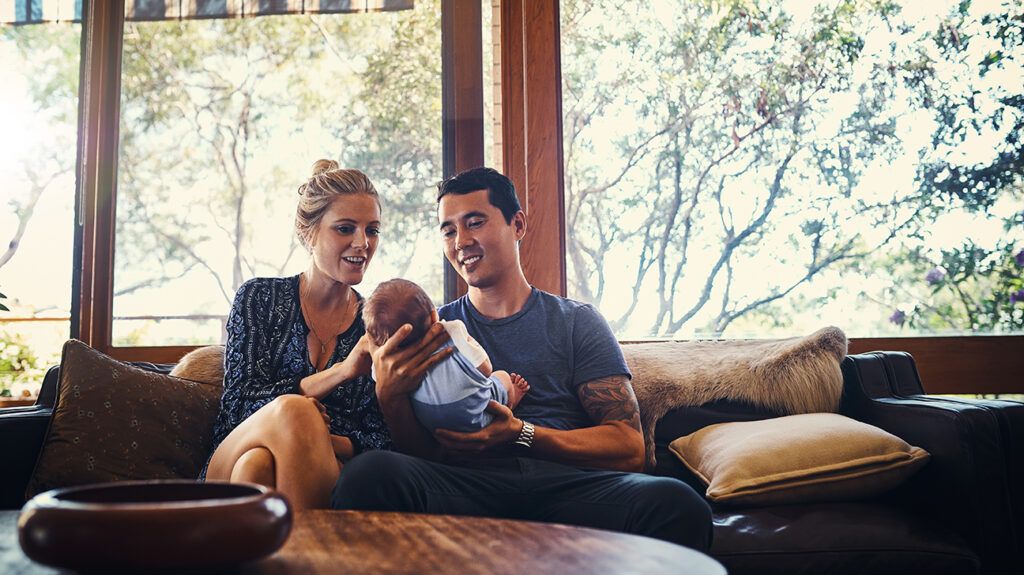 A father and mother, sitting on a couch in front of large windows holding a newborn baby