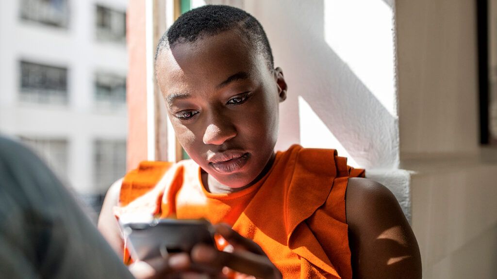 Black young woman in an orange top sitting by a window in the sunlight looking at her phone