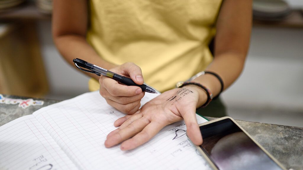A person sitting at a table taking notes from a book and writing them on their hand