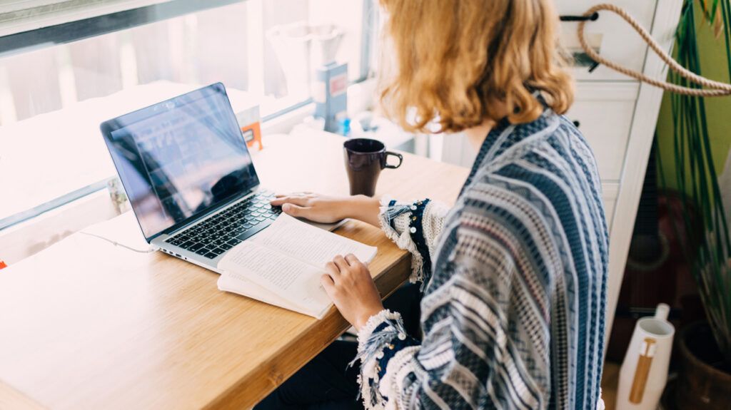 Person with long hair sitting at a desk looking at a laptop and book