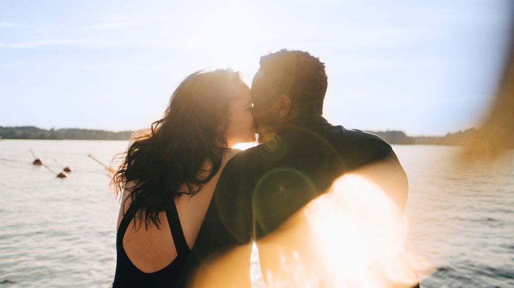 Couple viewed from behind, sitting on a beach and kissing by the water