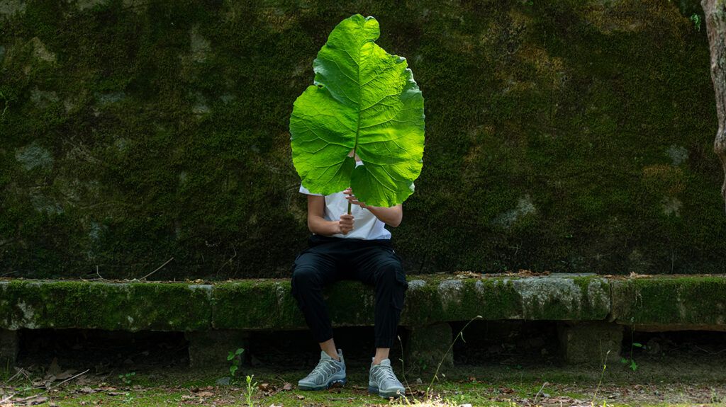 Person with autism sitting in the green outdoors, masking their face with a leaf