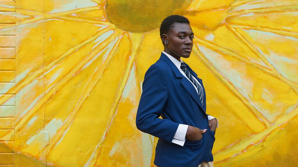 Androgynous Black woman in a suit standing near a yellow mural