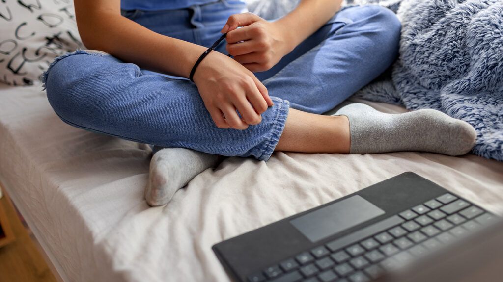 Teenager sitting cross-legged on a bed in front of a laptop; only their lower body is visible