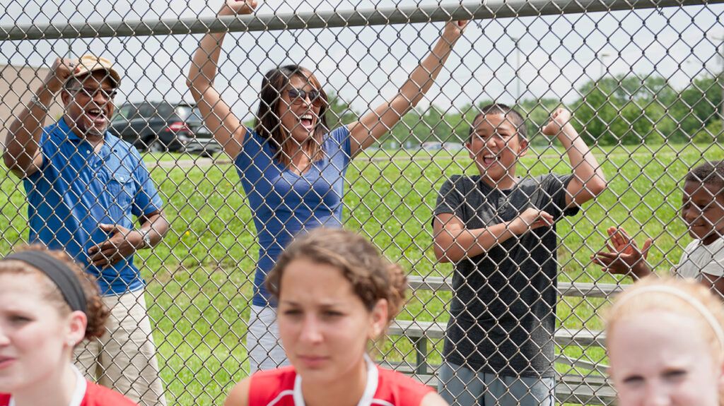 Family cheering for teen softball players behind fence