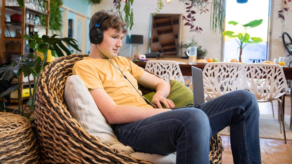 A teenage male sitting in a wicker chair with a laptop wearing headphones