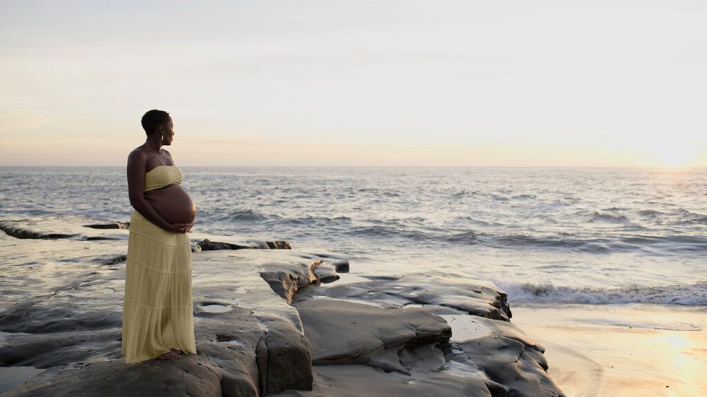 a pregnant woman standing on rocks at the beach looking into the distance